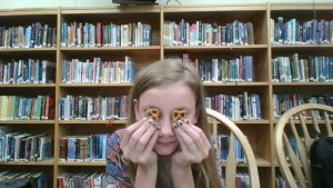 Girl with preztels covering her eyes, in front of bookshelf.